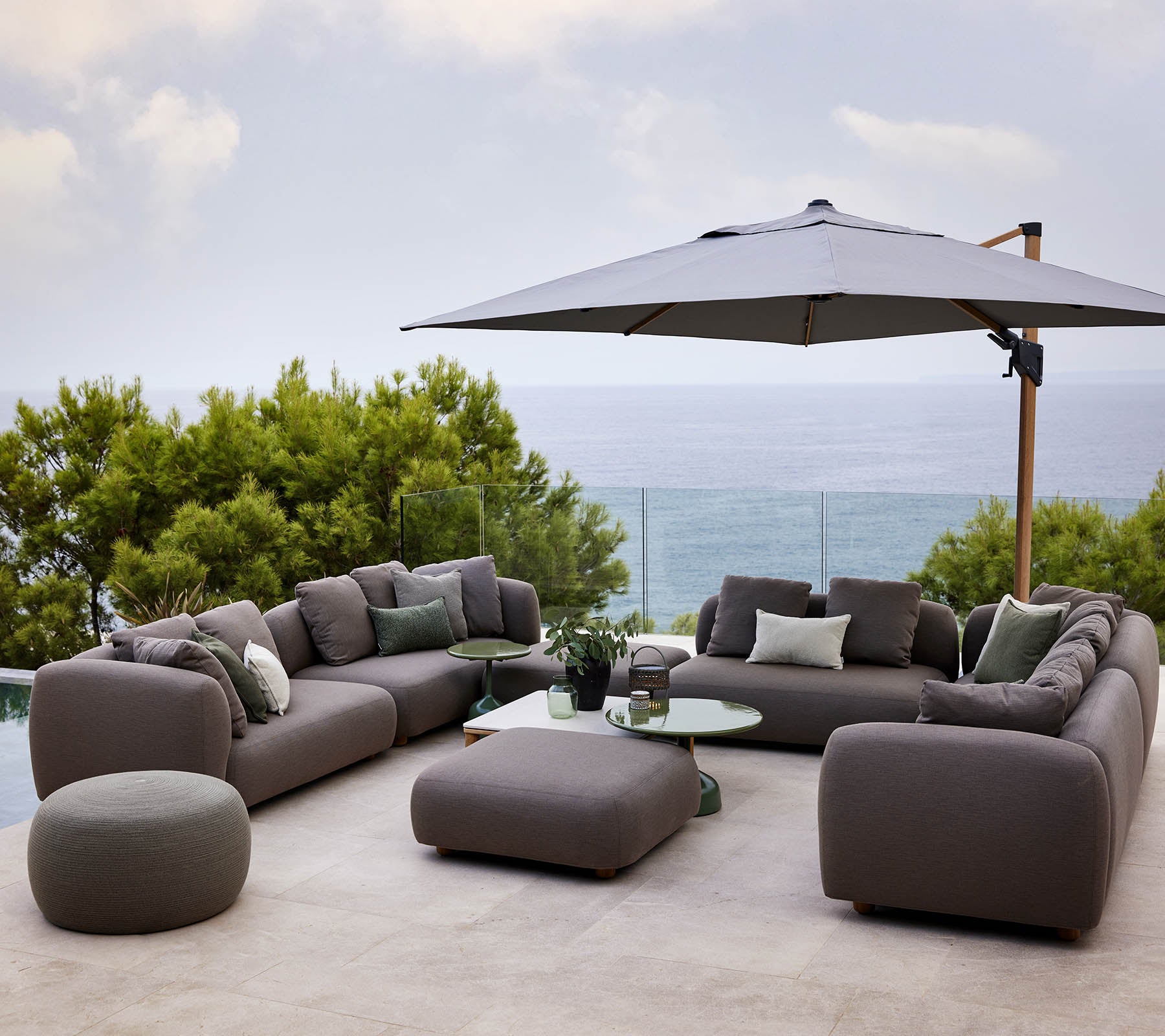 Modernes Loungemöbel-Set mit plüschigen Kissen, begleitet von einem runden Tisch und einem großen Sonnenschirm, umgeben von Grünpflanzen und einer malerischen Aussicht.