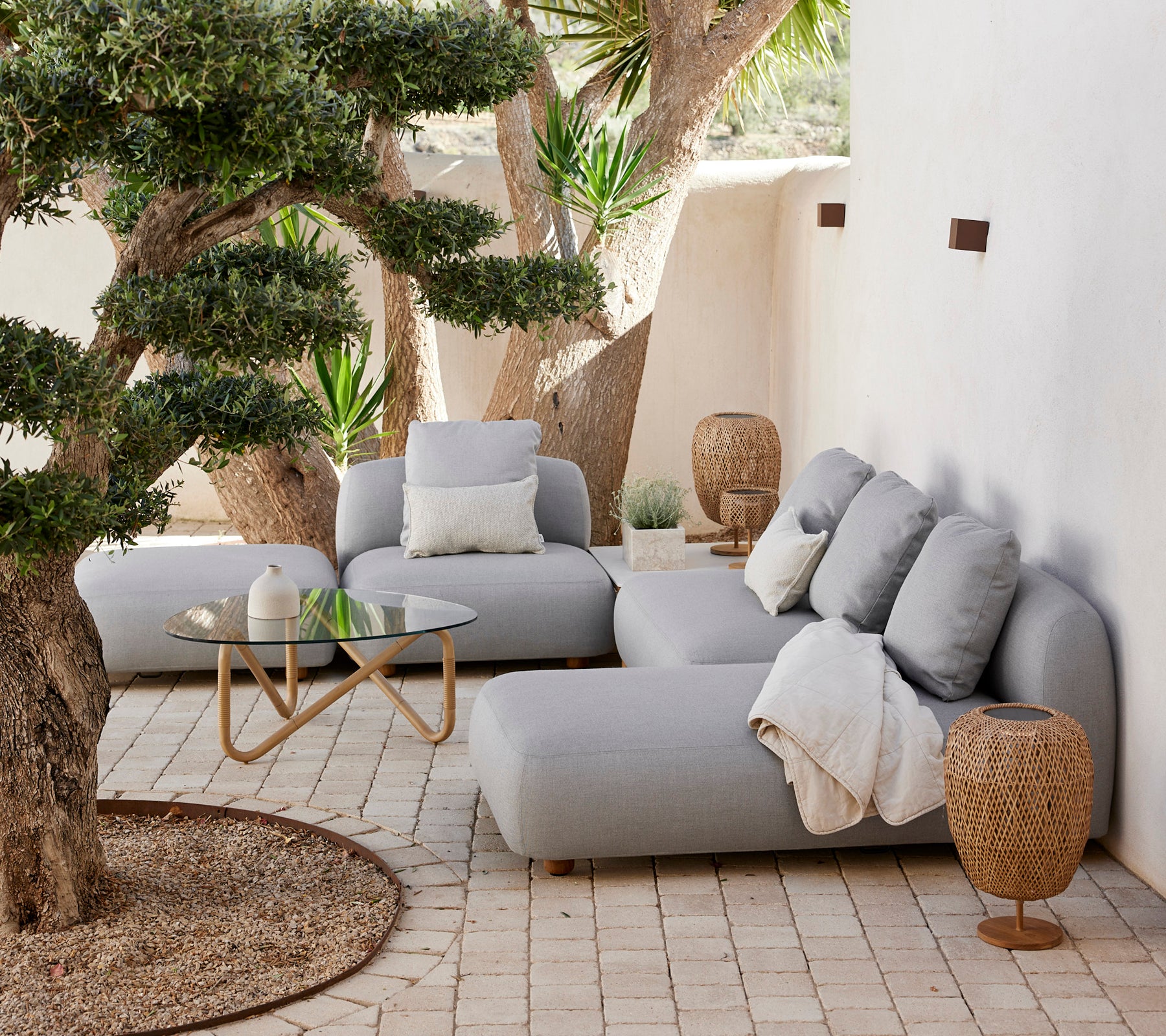 Modernes Lounge-Areal mit einem grauen Sofa, Kissen, einem Glastisch und einem geflochtenen Beistelltisch.