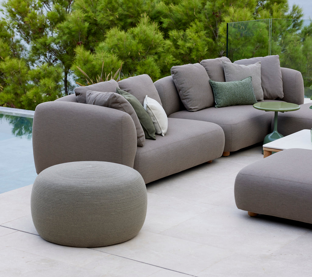 Großer taupefarbener runder Fußhocker neben einem modernen Sofa mit grünen Kissen, umgeben von üppigem Grün und einer friedlichen Atmosphäre.