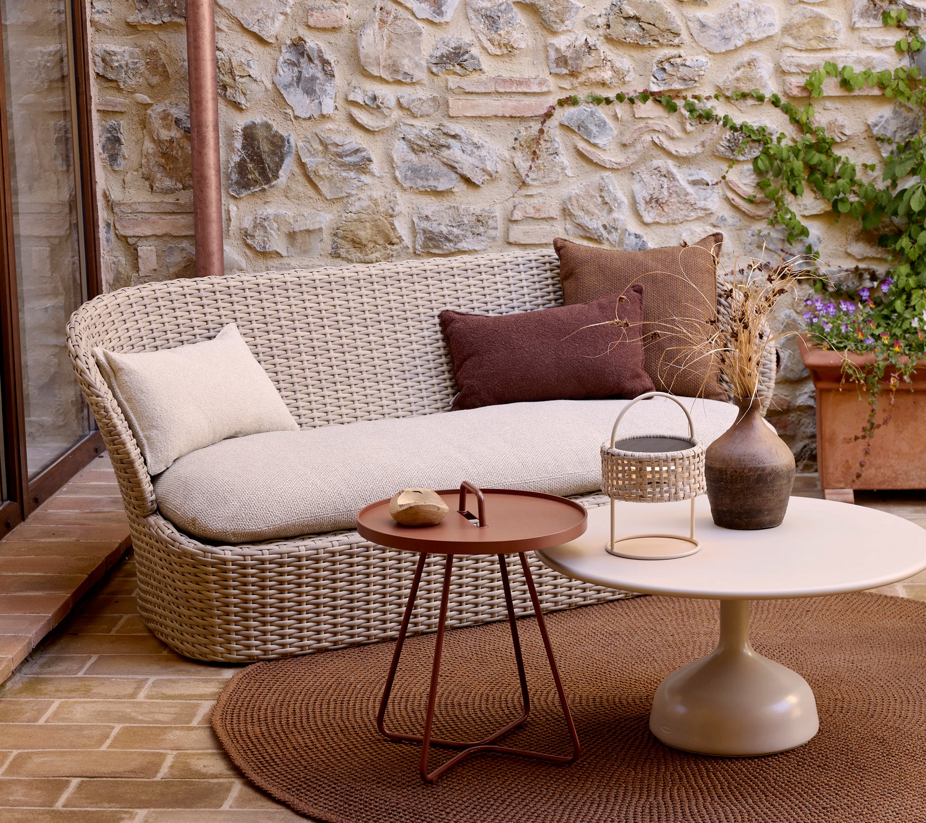 Ein modernes Zweisitzer-Sofa mit dekorativen Kissen, kombiniert mit einem runden Couchtisch und Pflanzen, vor einem Stein-Hintergrund.