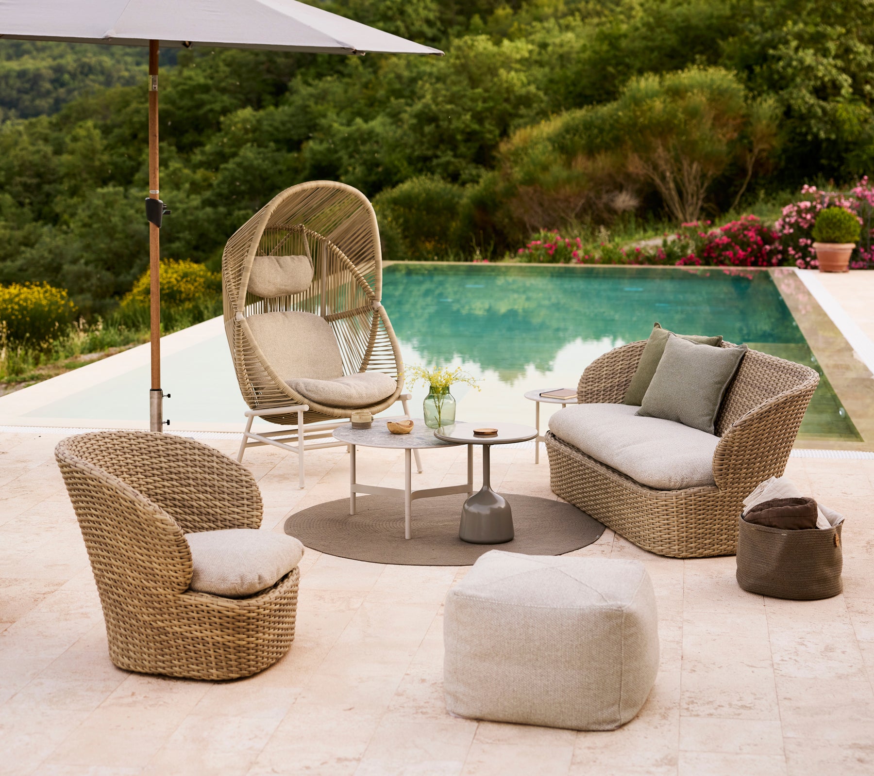 Küstenlounge-Setup mit einem Hive-Stuhl, zwei geschwungenen Stühlen, einem Divine-Hocker und einem kleinen Tisch neben einem Pool, umgeben von Grün.