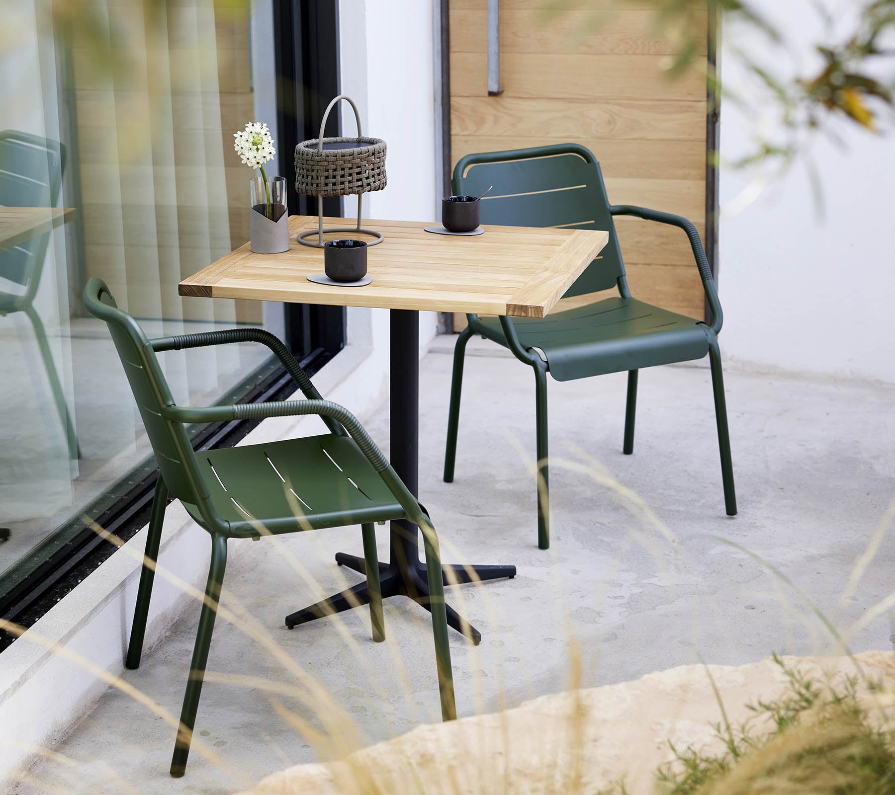 Eine minimalistische Einrichtung mit einem Holztisch und grünen Stühlen, die mit einem kleinen Blumengesteck und einer dekorativen Laterne versehen ist.