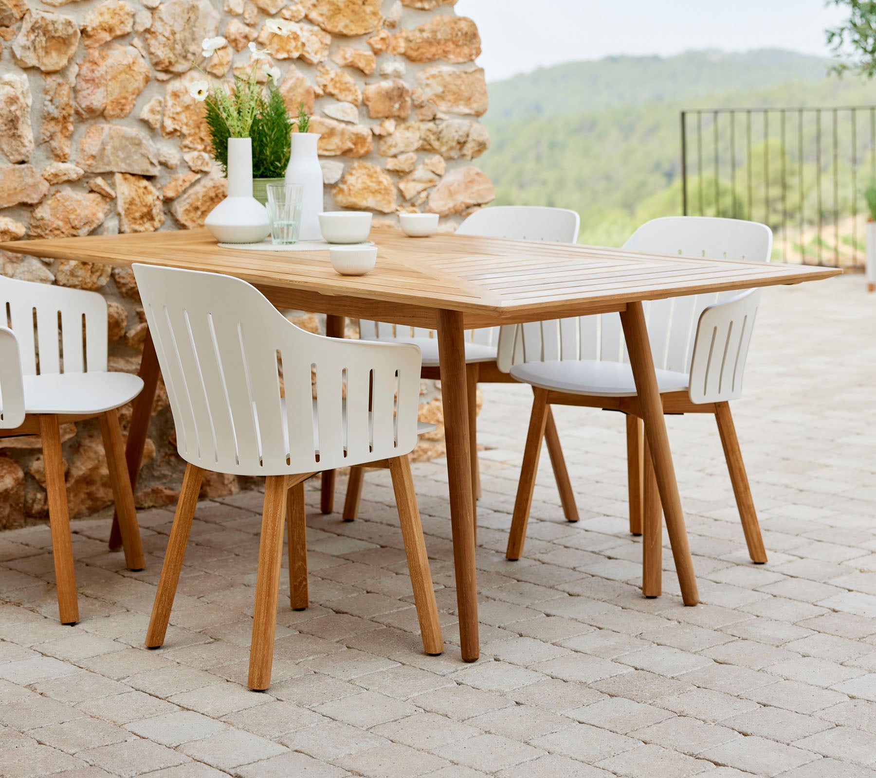 Ein Holztisch umgeben von weißen Stühlen, ergänzt durch dekorative Pflanzen und einfaches Geschirr, vor einem Hintergrund aus Naturstein.