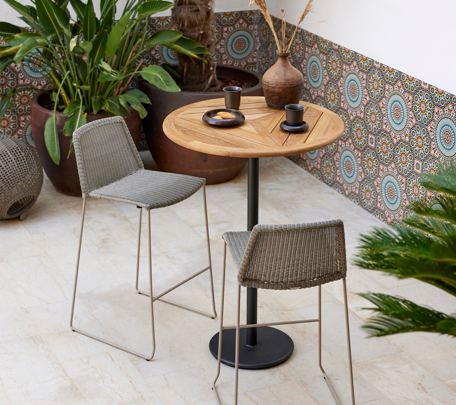 Ein stilvolles Bistro-Set mit einem runden Holztisch und zwei geflochtenen Hockern, begleitet von dekorativen Pflanzen und modernem Geschirr.
