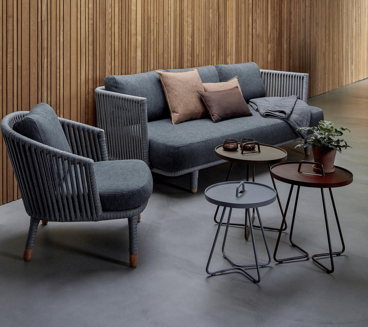 Stylishe Sitzanordnung mit einem grauen Sofa mit Kissen und zwei Akzentstühlen, begleitet von drei modernen Tischen und einer Topfpflanze.