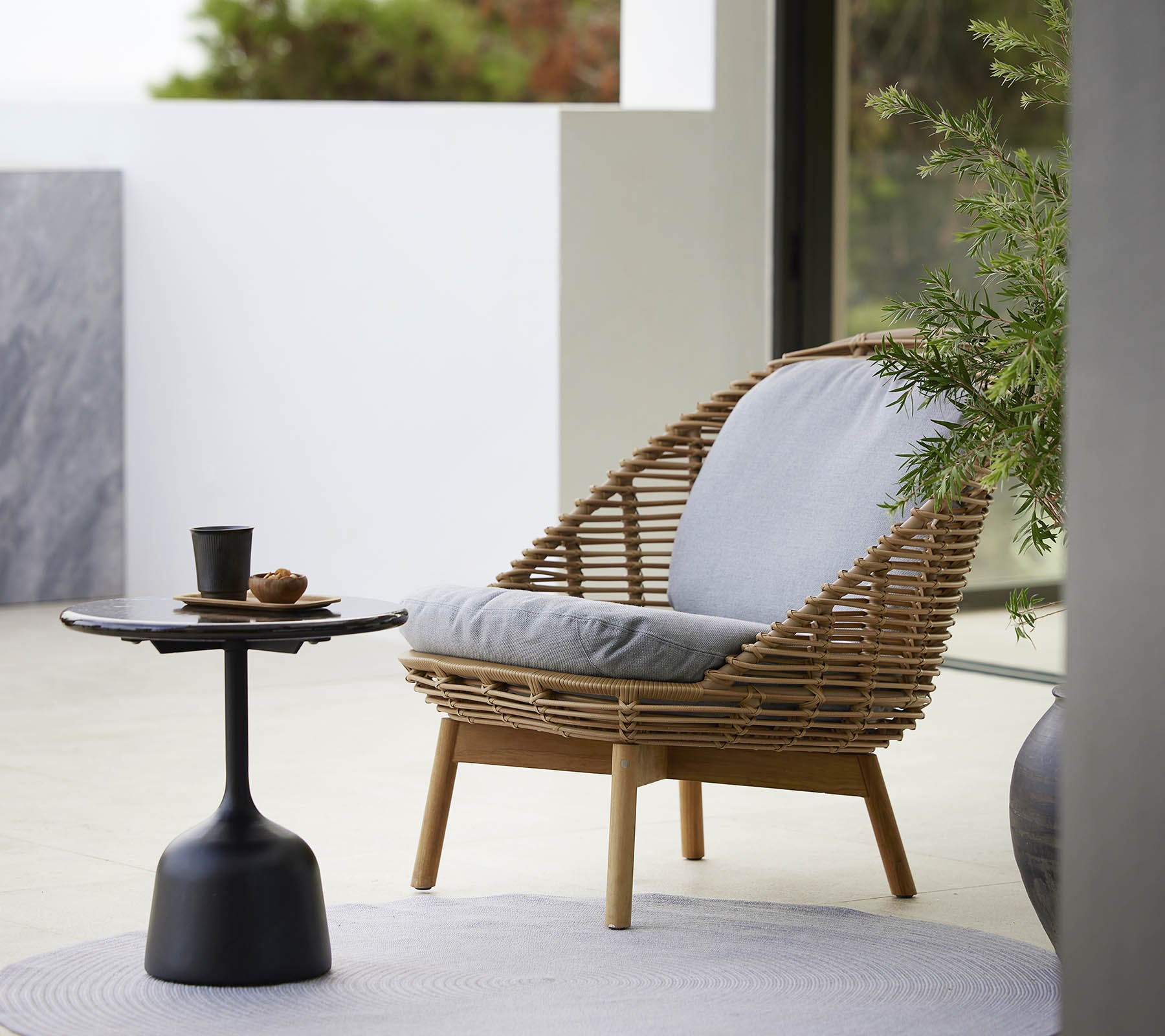 Ein stilvoller Lounge-Stuhl mit geflochtenem Design und weichen Kissen, begleitet von einem schlanken Beistelltisch und einer kleinen Pflanze. Ideal zum Entspannen.