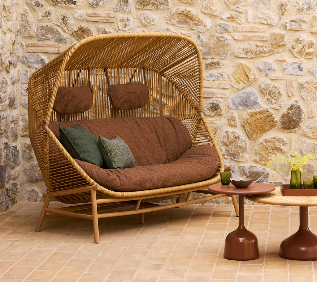 Eine stilvolle Rattan-Couch mit braunen Kissen und grünen Dekokissen, begleitet von zwei modernen Couchtischen.