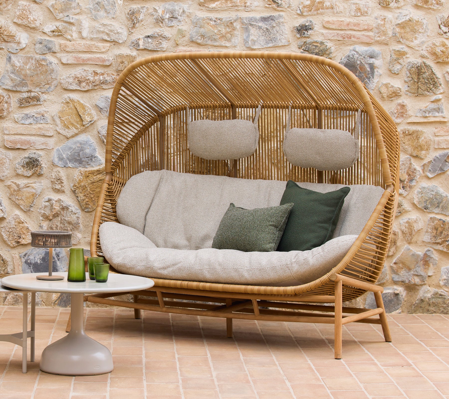 Hive Sofa mit plüschigen Kissen und grünen Akzenten, begleitet von Glaze Couchtischen, vor einem natürlichen Stein Hintergrund.