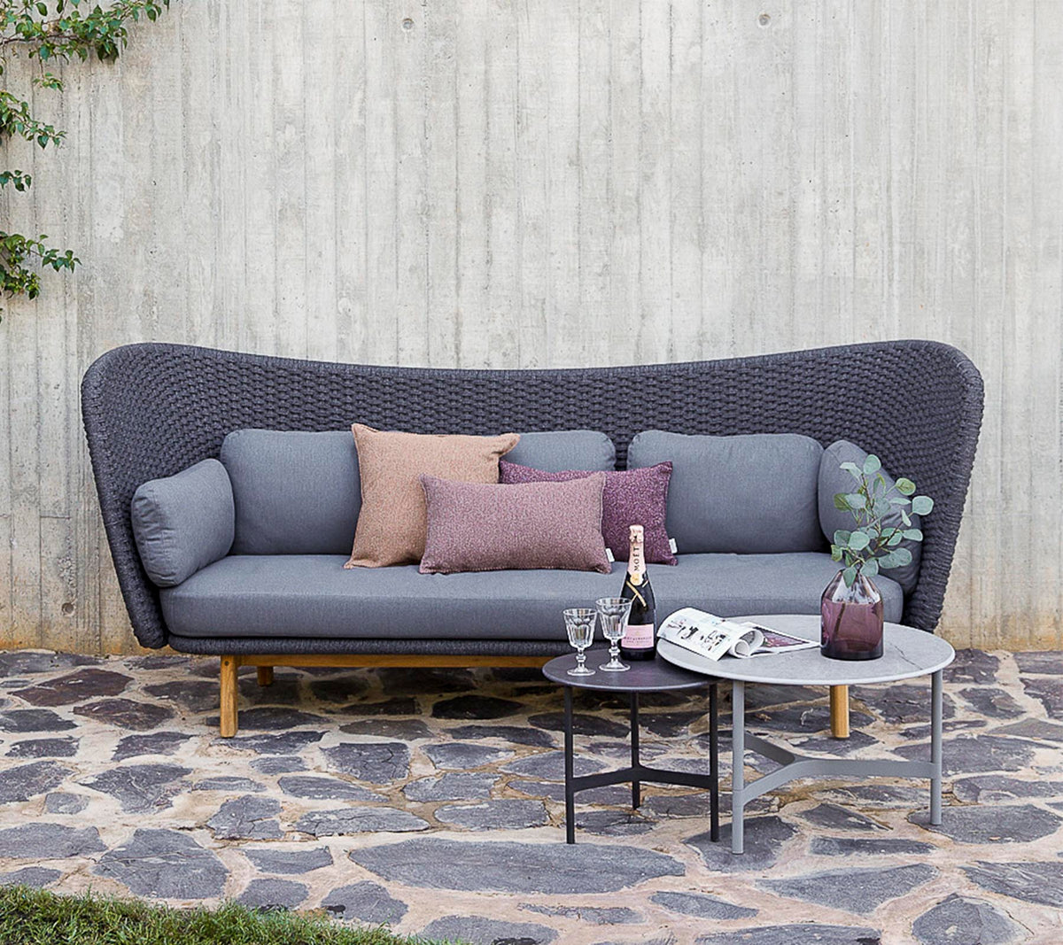 Ein modernes Sofa in Grau mit dekorativen Kissen, begleitet von einem kleinen Tisch mit Getränken und einer Vase, vor einer strukturierten Wand platziert.