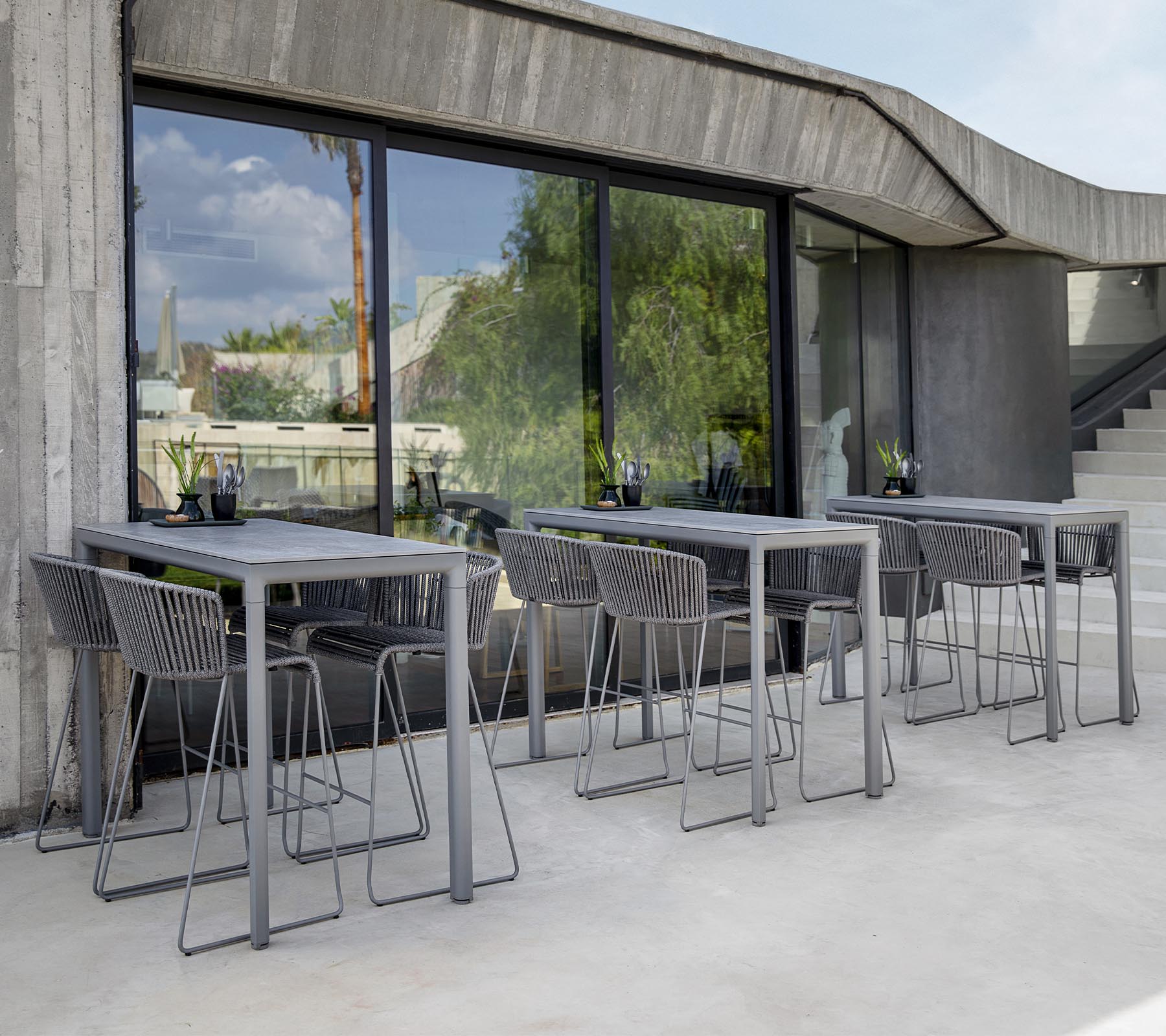 Moderne Sitzbereich mit grauen Tischen und Stühlen, großen Fenstern, die das Grün draußen widerspiegeln. Minimalistisches Design ästhetik.