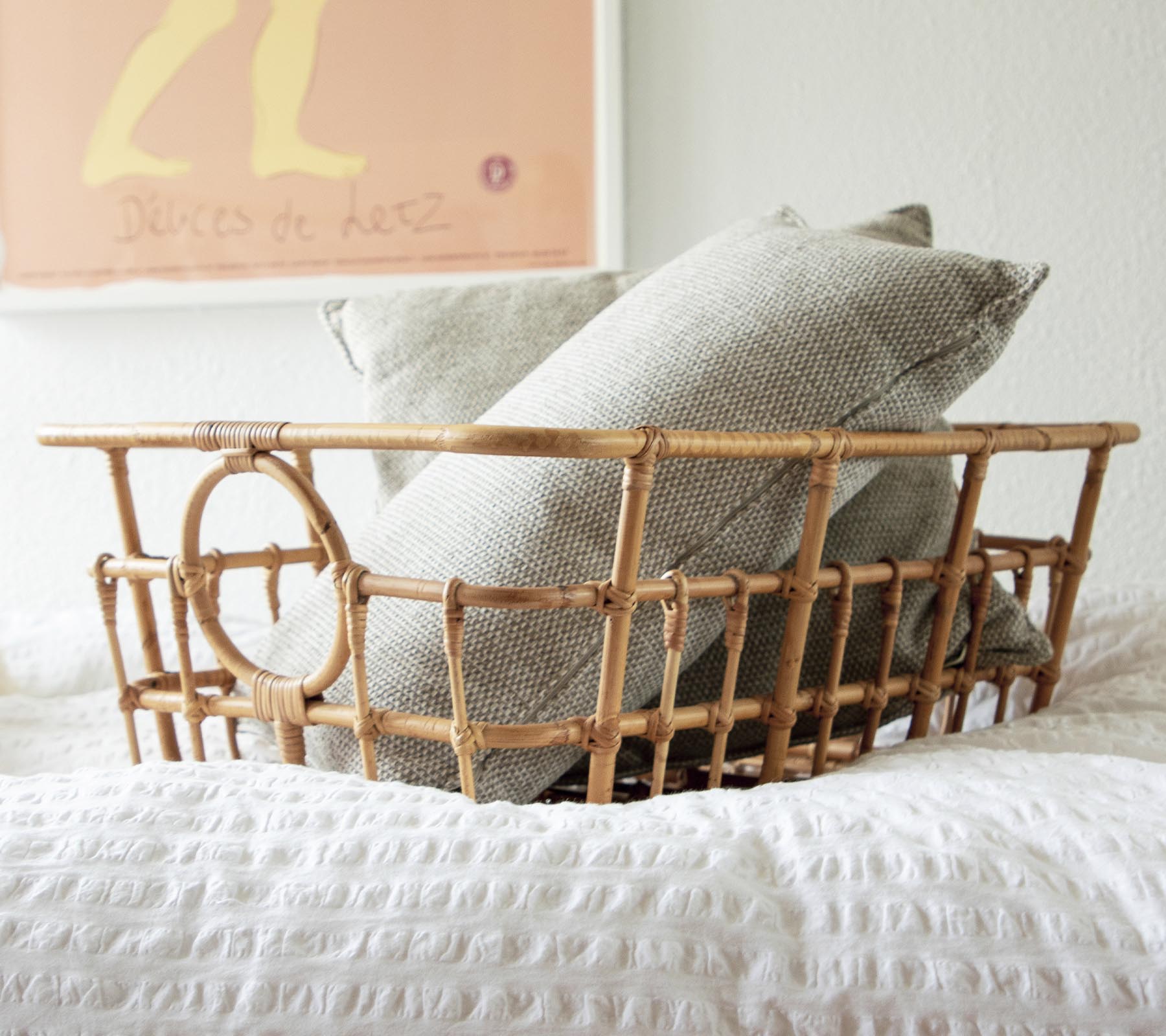 Rattan-Korb mit grauen und weißen Kissen in einem gemütlichen Schlafzimmer-Ambiente.