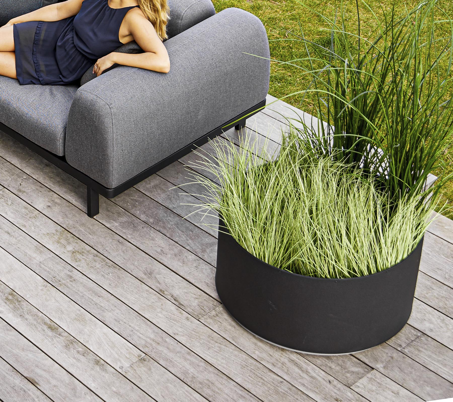 Ein stilvolles graues Sofa neben einem runden schwarzen Blumentopf, gefüllt mit hohem grünen Gras, auf Holzterrasse gesetzt.