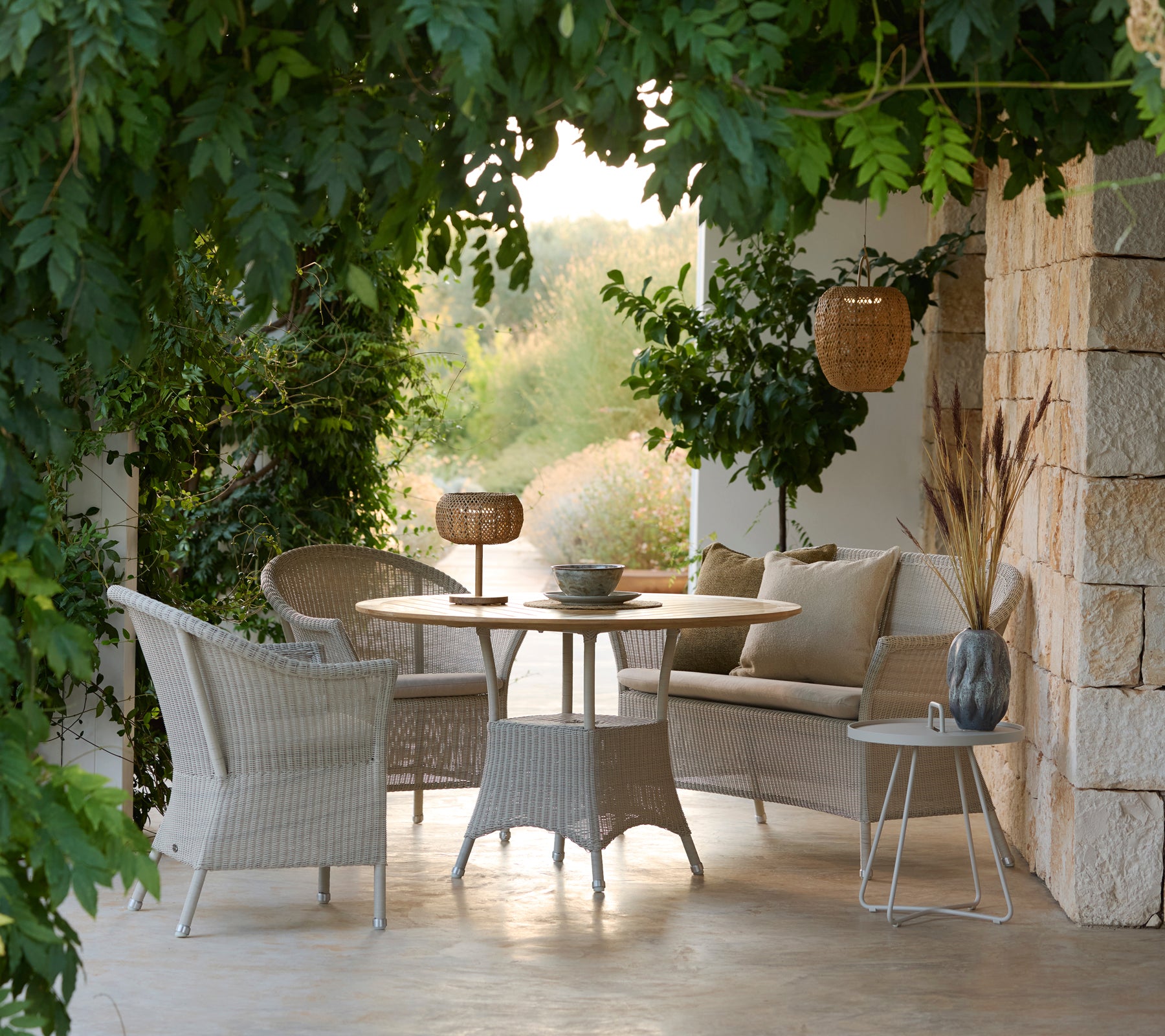 Zeitgemäßes Loungemöbel-Set mit geflochtenen Stühlen, rundem Tisch und Kissen in einer ruhigen Außenumgebung.