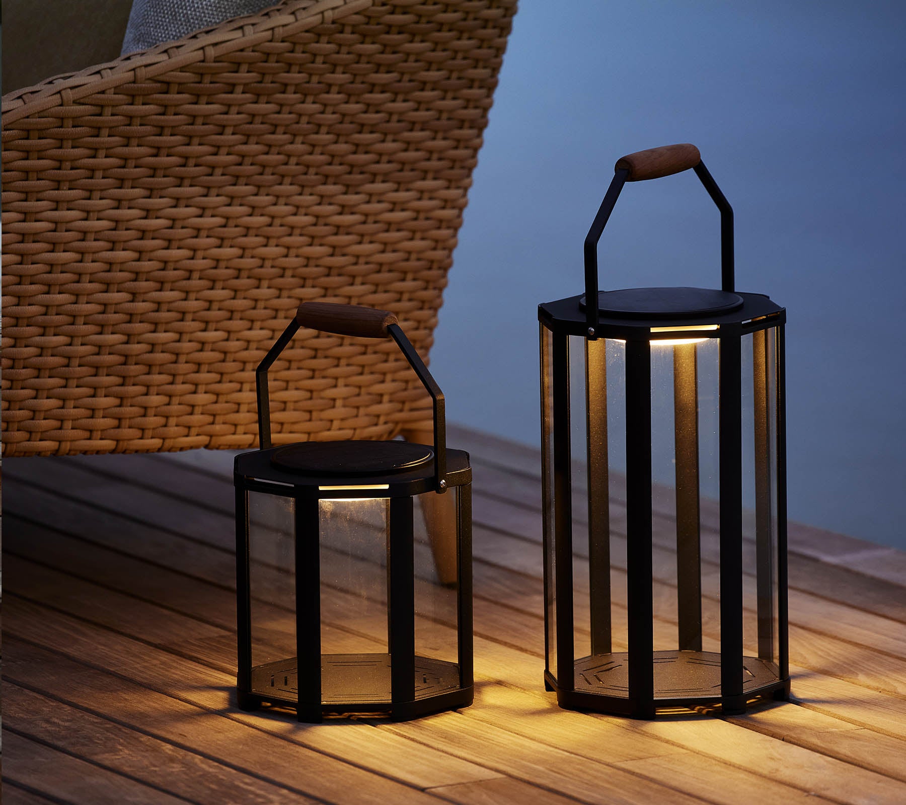 Zwei stilvolle Lightluxe Laternen mit Glaspanelen und Griffen, die ein warmes Licht ausstrahlen, neben einem geflochtenen Stuhl platziert.