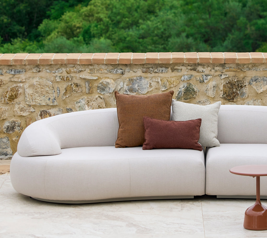 Ein geschwungenes Sofa, geschmückt mit Rise-Kissen in erdigen Farben, neben einem stilvollen Glaze-Beistelltisch, vor einem steinernen Hintergrund.