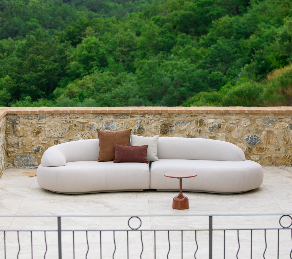 Ein modernes, geschwungenes Sofa mit neutralem Bezug und dekorativen Kissen, ergänzt durch einen eleganten Beistelltisch in einer natürlichen Umgebung.