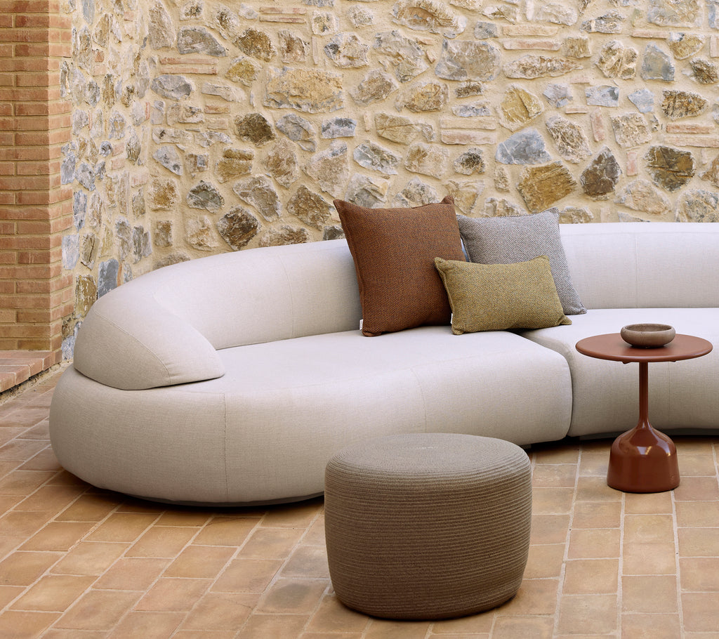 Kurven sofa mit dekorativen Kissen, einem runden Hocker und einem glasierten Beistelltisch auf einem strukturierten Boden mit einer Steinwand im Hintergrund.