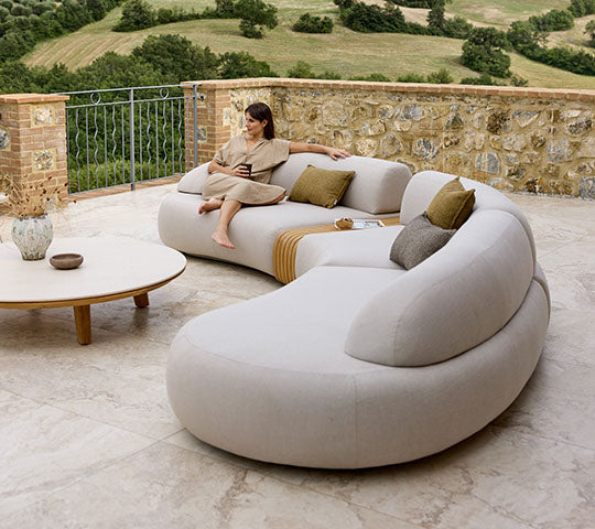 Ein sanft geschwungenes Sofa mit Kissen und einem Holztisch, begleitet von einer Person, die bequem sitzt und die Aussicht genießt.