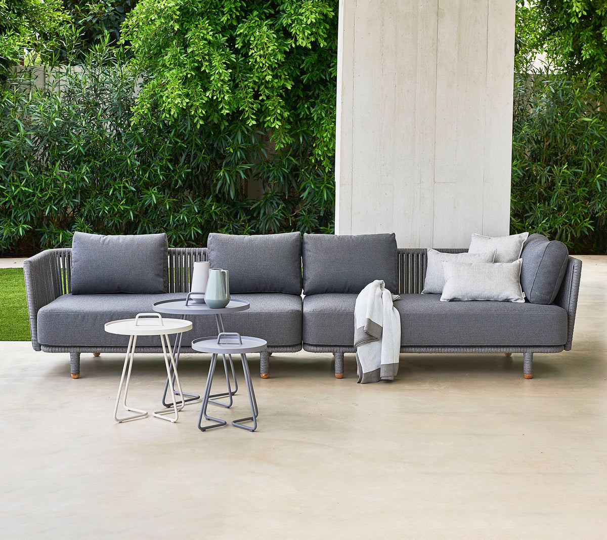 Moderne graues Ecksofa mit Kissen, begleitet von zwei stilvollen Beistelltischen, vor einem Hintergrund aus üppigem Grün.