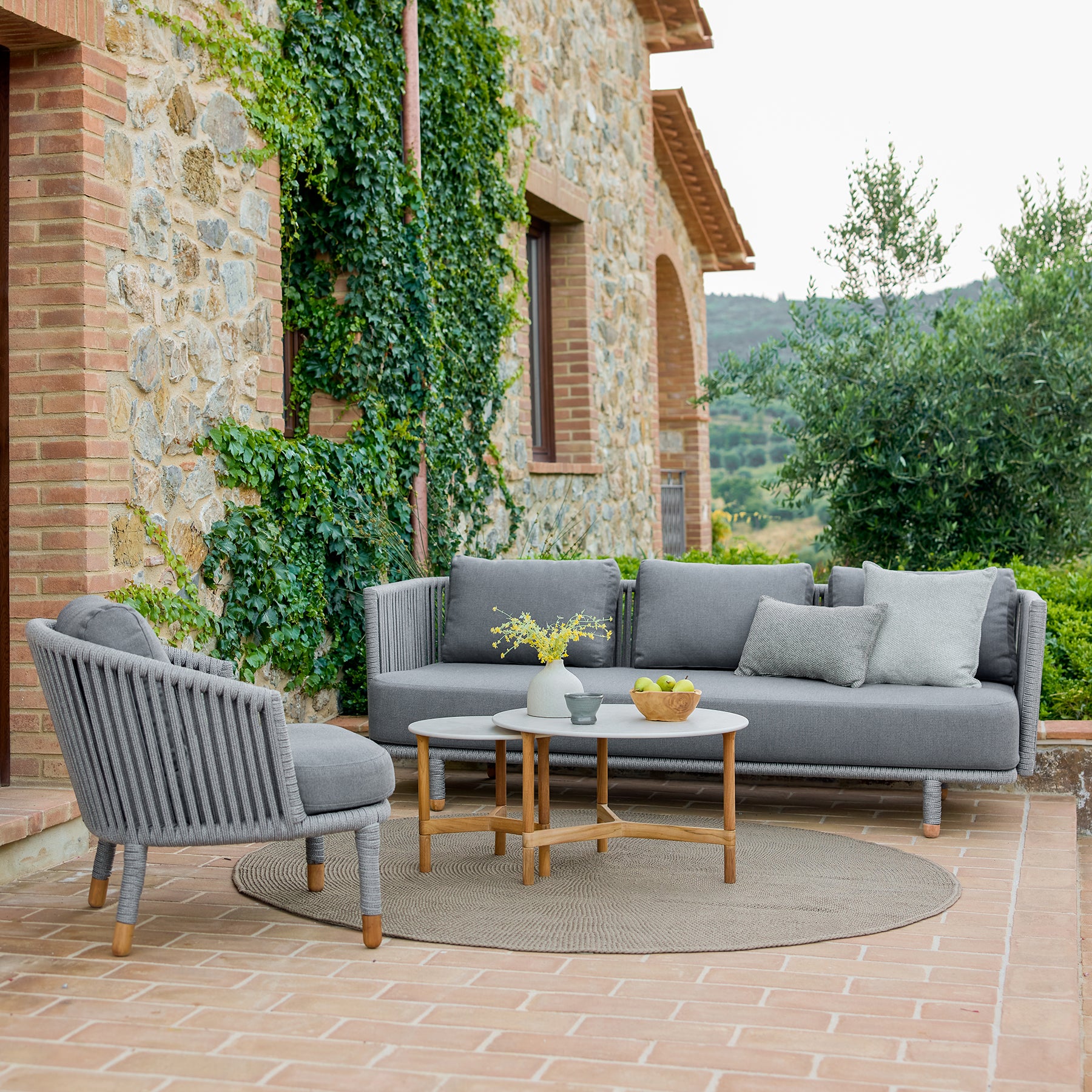 Modernes graues Sofaset mit passendem Sessel und einem runden Couchtisch, umgeben von Grün und rustikaler Steinearchitektur.