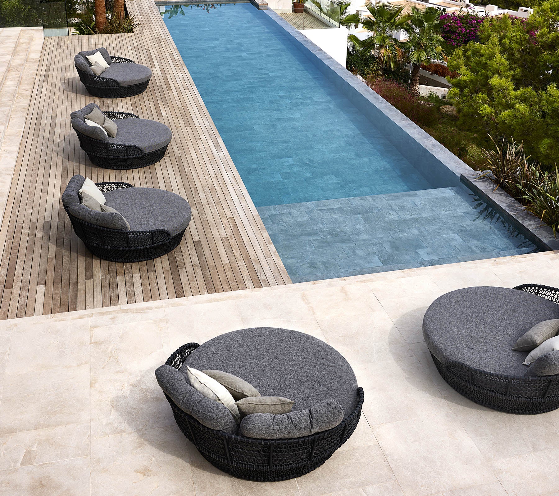 Vier runde Liegen am Meer mit Kissen sind neben einem eleganten Swimmingpool angeordnet, der von Grünflächen und Holzdecks umgeben ist.