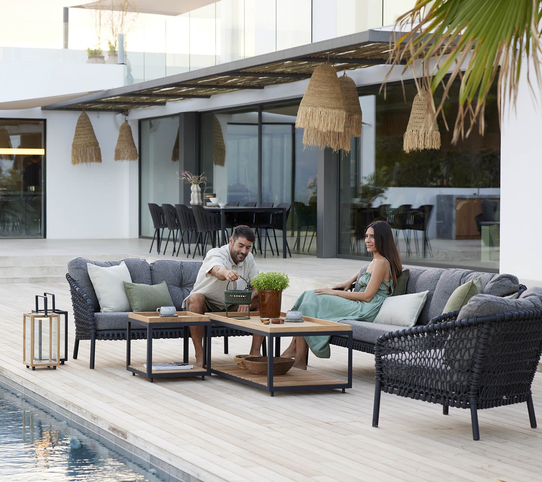Paar genießt Freizeit auf einer gemütlichen Lounge-Garnitur, umgeben von grünen Pflanzen und stilvollem Dekor, mit einem ruhigen Schwimmbecken in der Nähe.