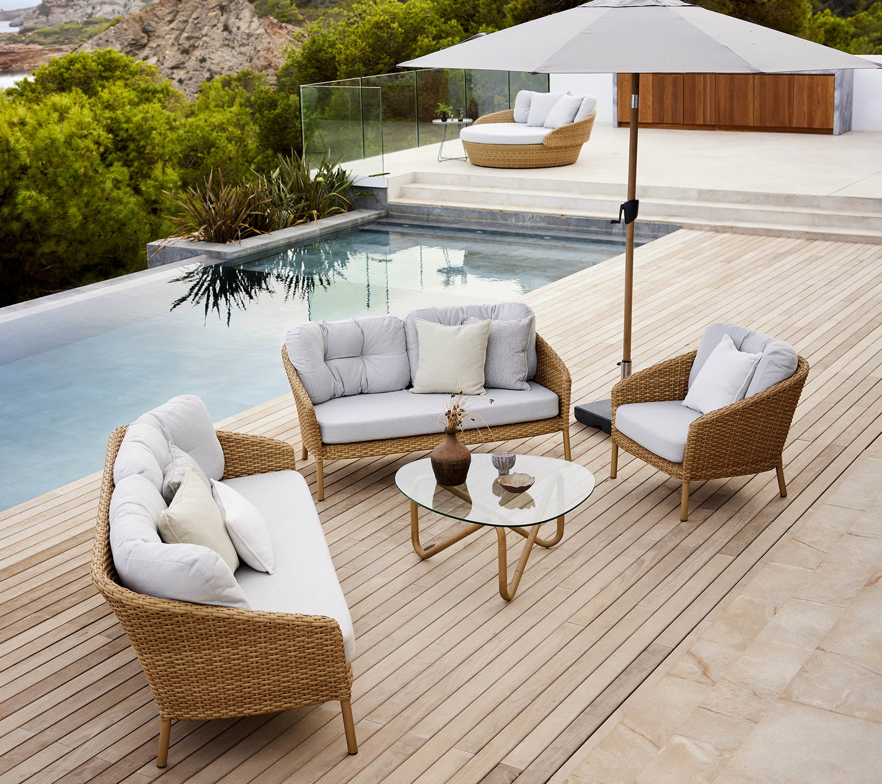 Moderne Außenlounge-Bereich mit einem Glastisch, bequemen Sitzgelegenheiten und einem Sonnenschirm am Poolrand mit üppigem Grün.