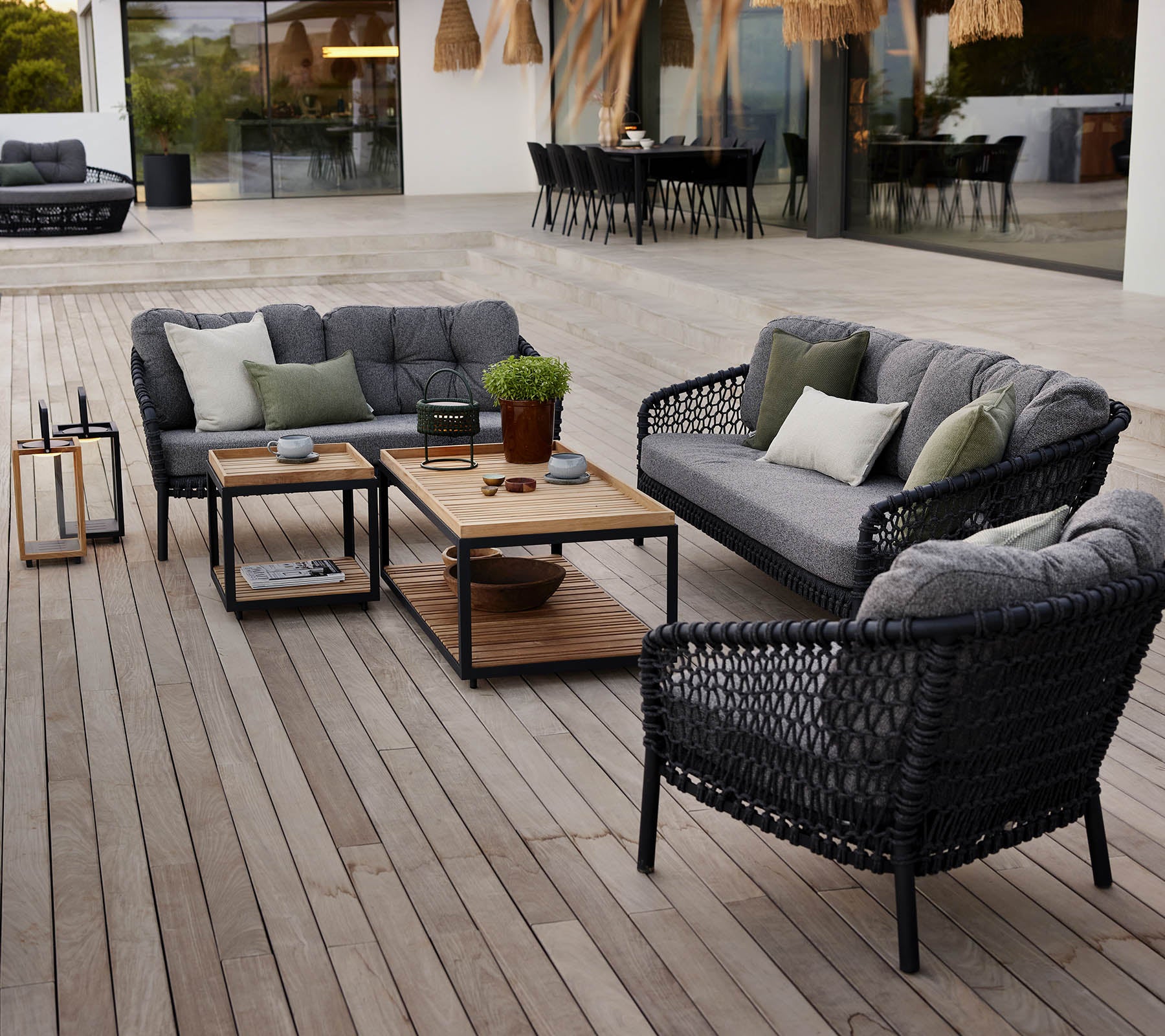 Stylischer Außenbereich mit schwarzen Rattanmöbeln, grauen Kissen, einem Holztisch und Topfpflanzen auf einer Holzterrasse.