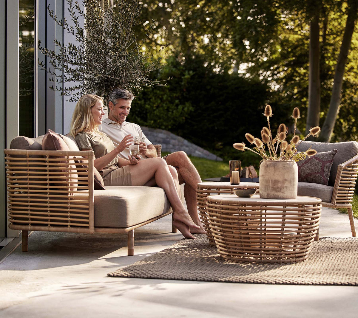 Ein Paar genießt Getränke auf einem Sense-Sofa und einem Loungesessel, begleitet von einem stilvollen Korbkaffee-Tisch und einer dekorativen Vase.