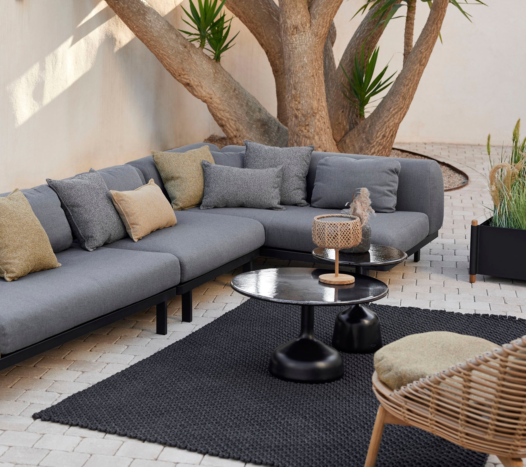 Loungebereich mit einem grauen modularen Sofa mit dekorativen Kissen, einem eleganten Couchtisch und einem strukturierten Teppich, umgeben von Grün.