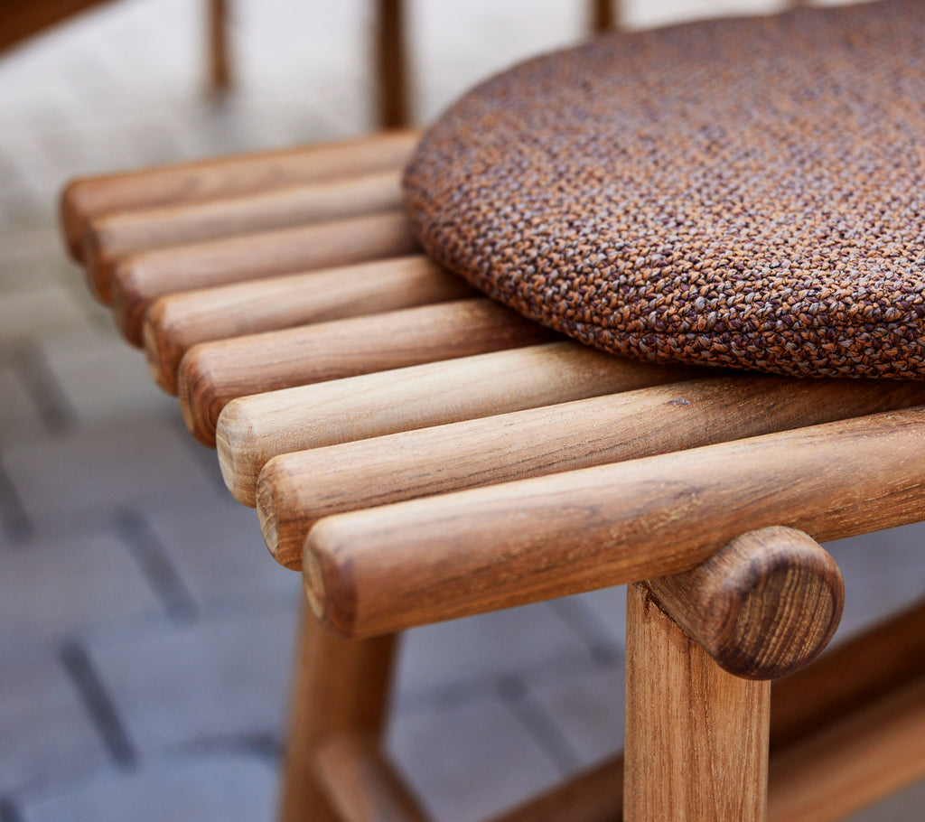 Detailaufnahme einer Teakbank mit Holzstöcken und einem strukturierten runden Kissen oben, das Handwerkskunst und natürliche Materialien hervorhebt.