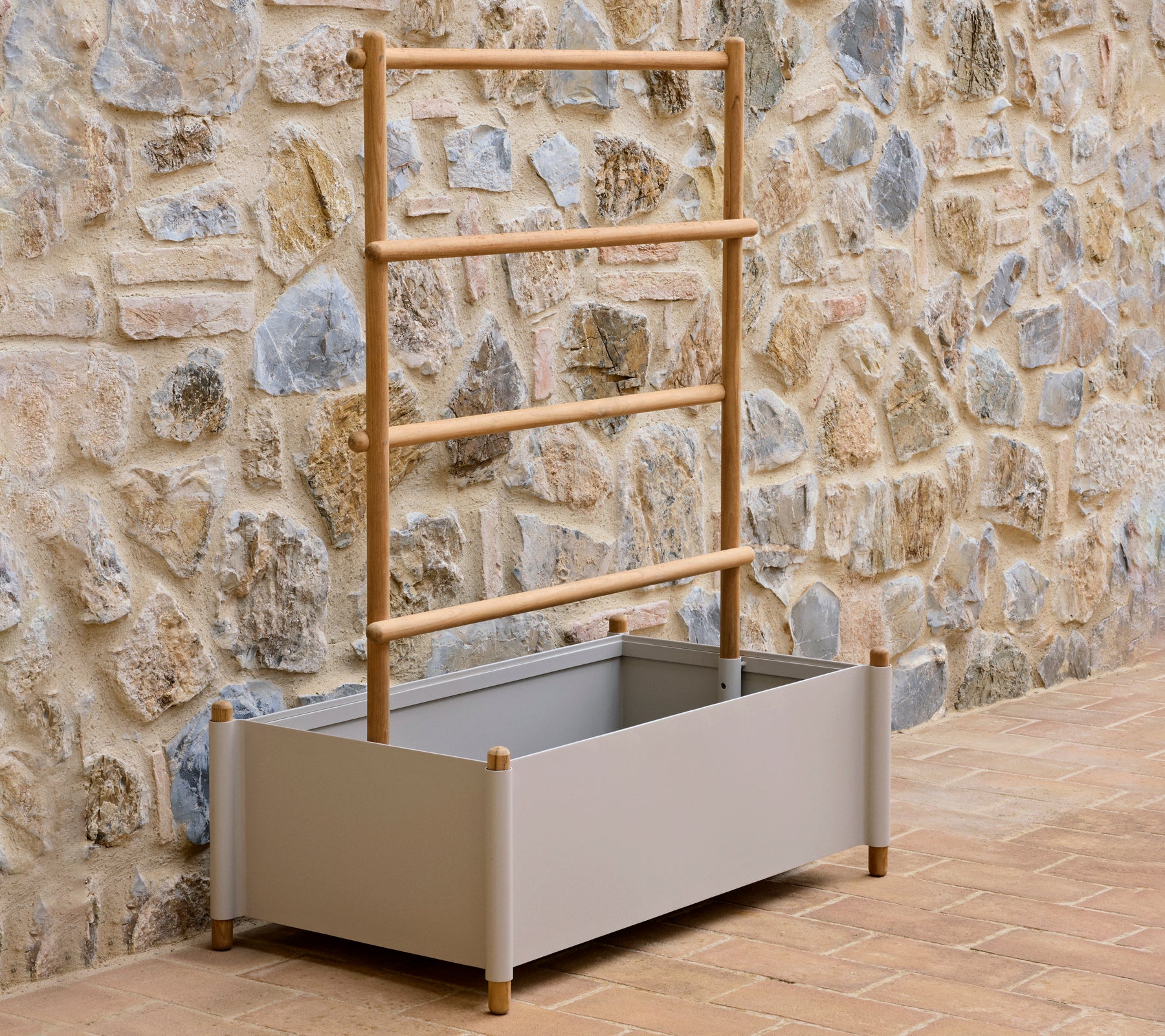 Ein grauer Aluminium-Pflanzkübel mit einer Holzleiter, designed für den Anbau von Pflanzen, vor einer strukturierten Steinmauer.
