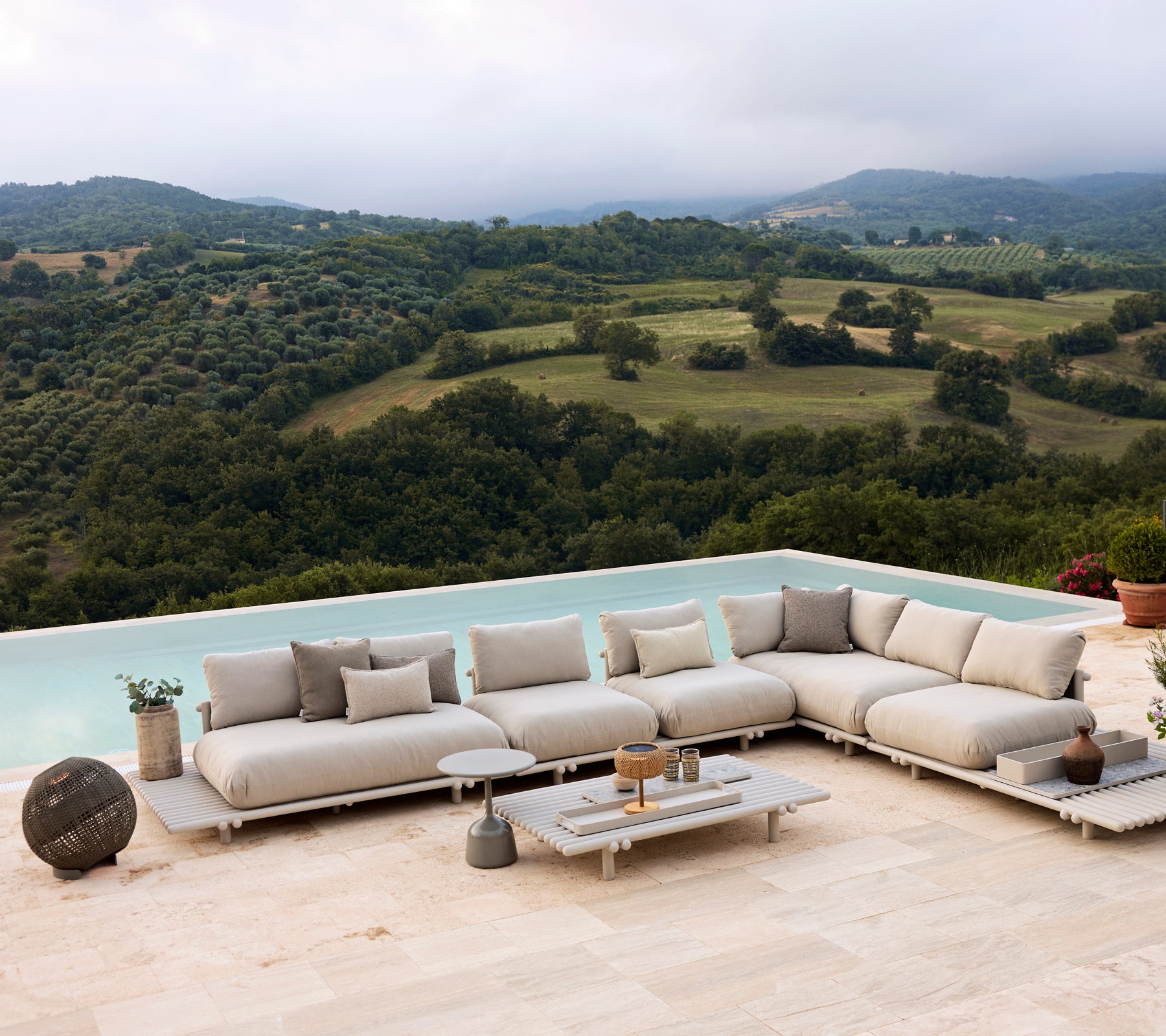 Aluminium Lounge-Sitzmöbel auf einer Plattform, mit neutralen Kissen und einer Illusion-Rundlampe, vor einer malerischen Landschaft.