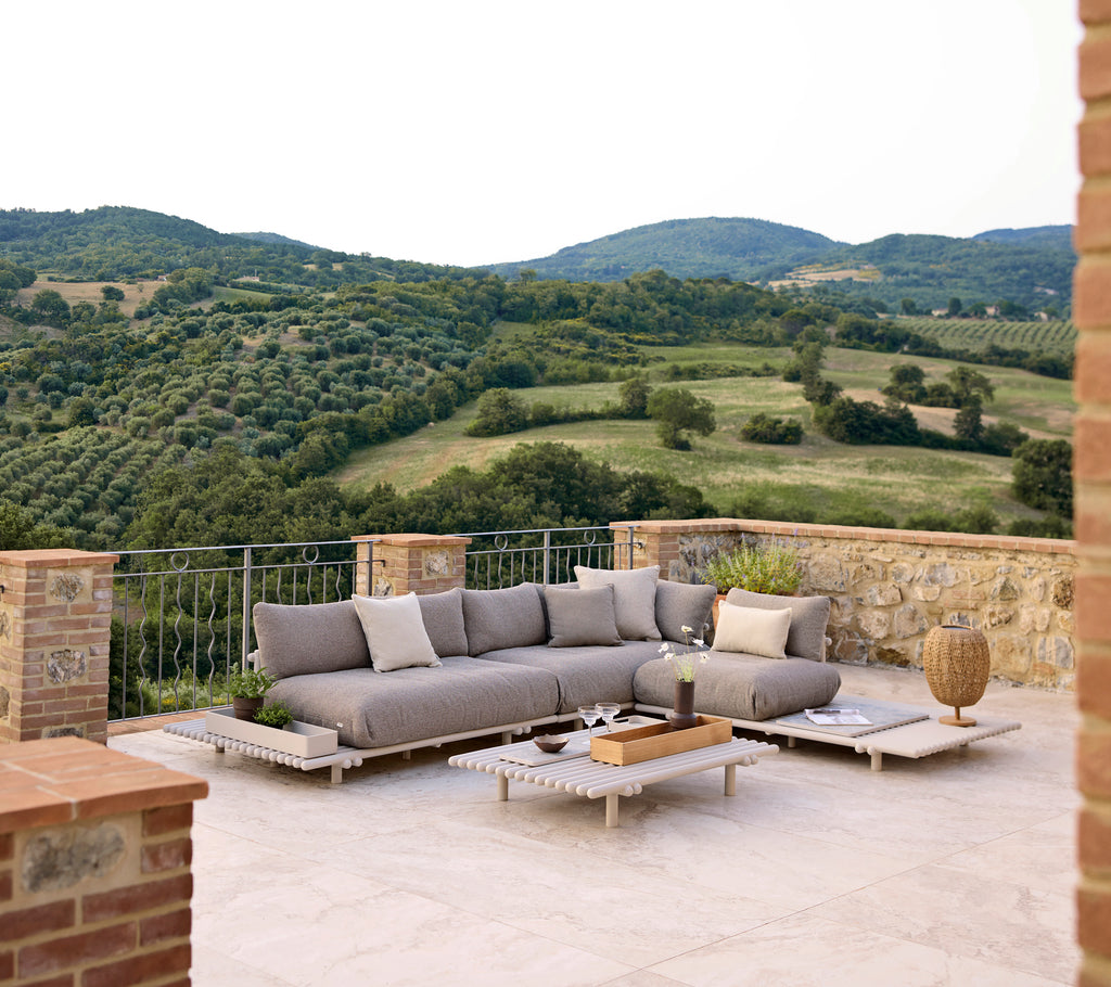 Stylischer Lounge-Bereich mit einem grauen Ecksofa und einem Holztisch, umgeben von üppigen Hügeln und einer malerischen Landschaft.