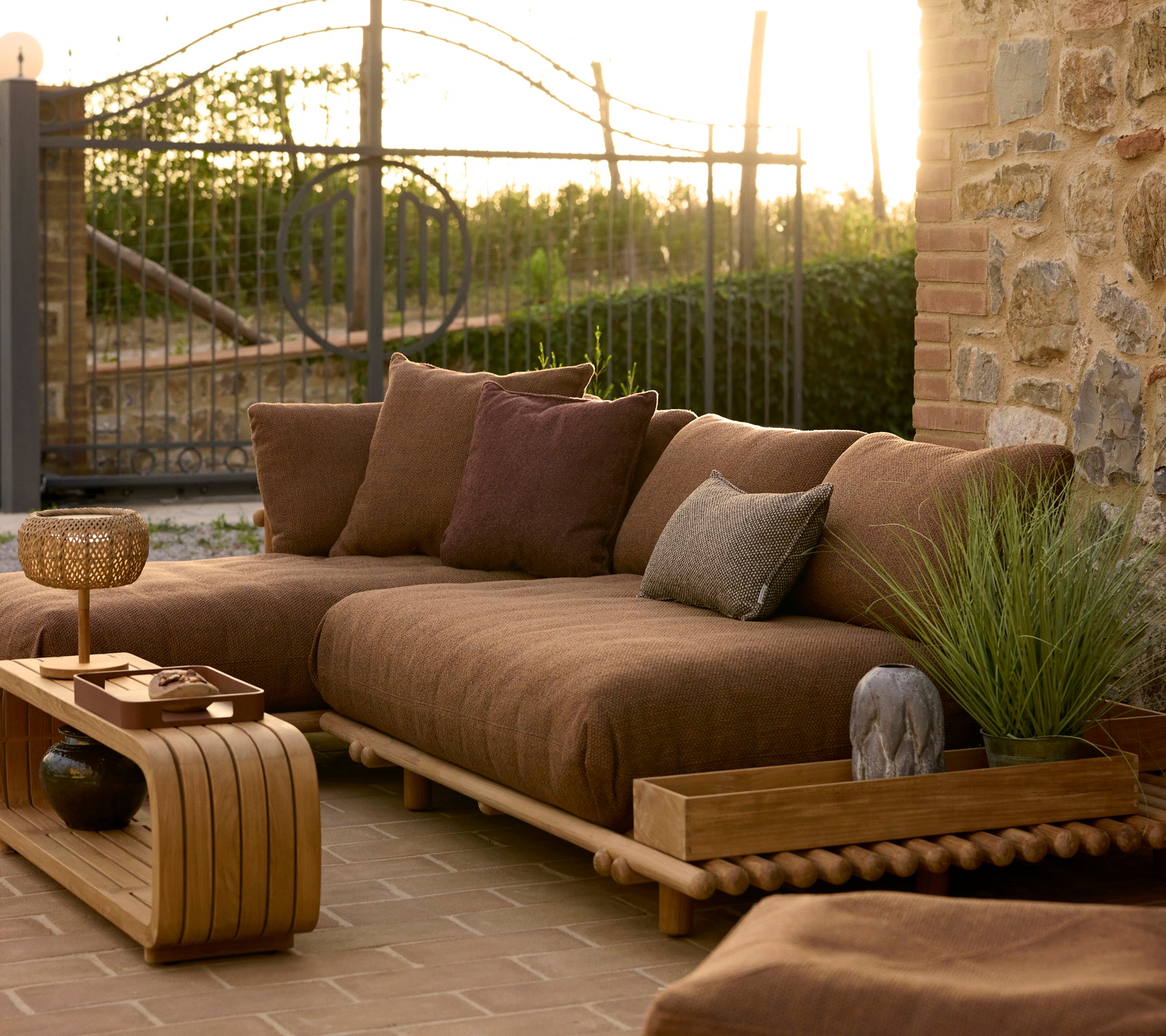 Lässiger Couchtisch und Teakholz-Club-Aufbewahrungskiste neben einer gemütlichen Sitzecke im Freien mit Kissen und Pflanzen im warmen Abendlicht.