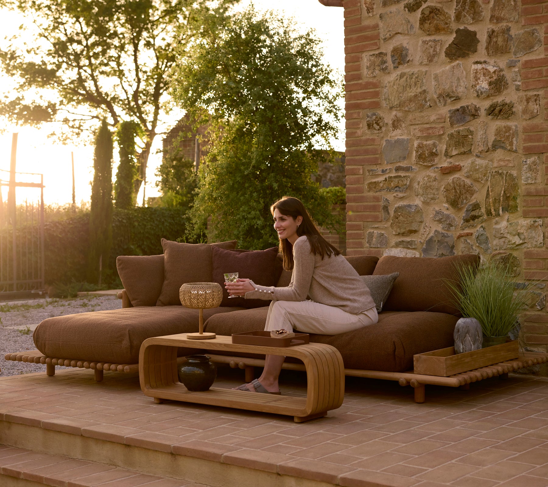 Eine Frau entspannt sich auf einem modernen Teak-Outdoor-Sofa mit einem sanften Couchtisch und einem dekorativen Tablett, vor einer steinernen Kulisse.
