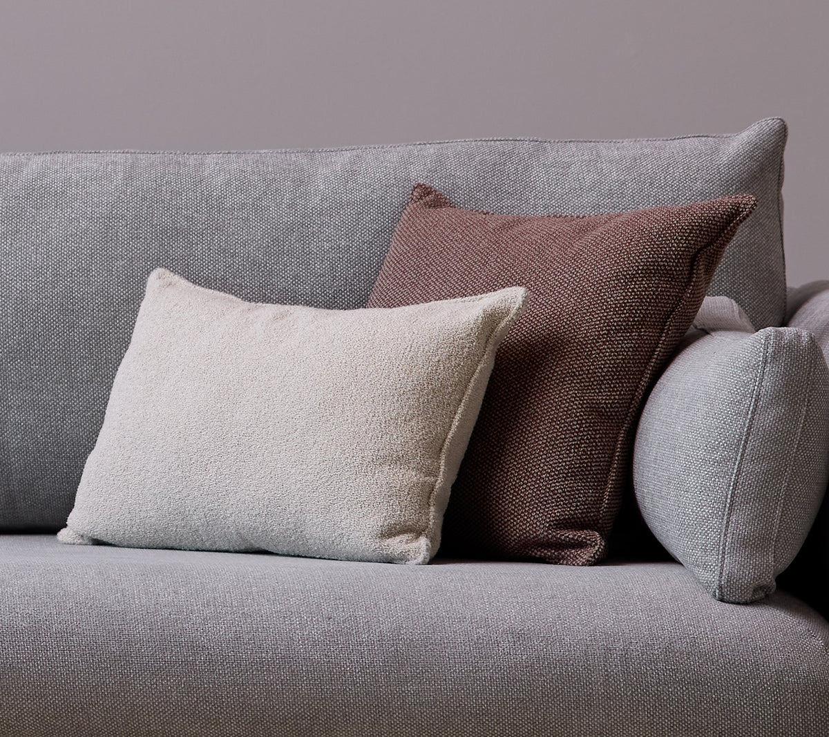 Ein weißes Scent-Kissenscatter und ein Wove dunkelbordeaux Kissen sind auf einem grauen Sofa platziert.